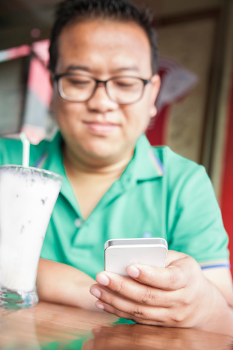 Indian man from Nagaland, India using his smart phone to text a friend at an outdoor cafe.  He wears a green collared shirt and glasses and a slight smile.  He is drinking a cold drink in the hot Indian summer season. Portrait-style image of this man who is in his late 20s.   Focus on the phone.