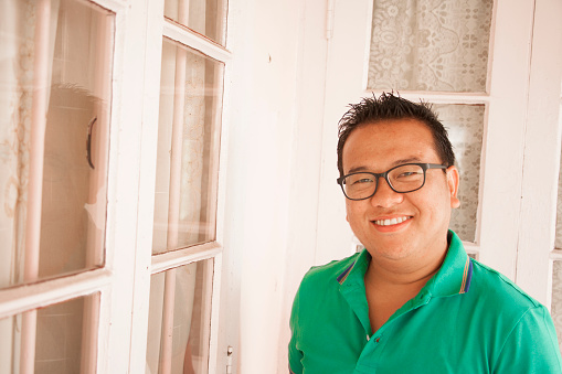 Indian man from Nagaland, India standing on his front porch at home.  He wears a green collared shirt and glasses and a slight smile.  He stands near a wall of outdoor windows. Portrait-style image of this man who is in his late 20s.  