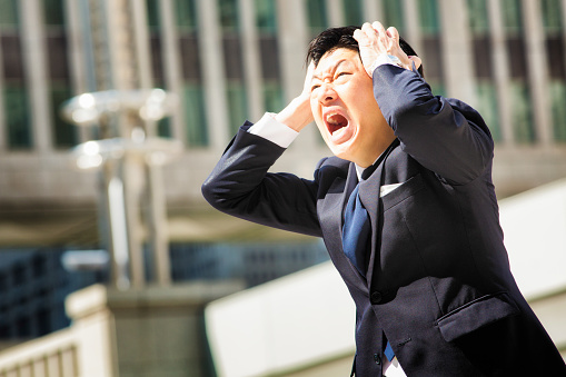 Furious angry Japanese office worker screams in despair while holding his head with both hands. Office building in the background. Photographed in Shinjuku, Tokyo, Japan.