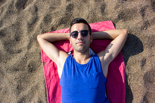 Relaxed young man lying down on towel on beach. He is resting on the towel. Young man wearing casual clothes. High angle view,horizontal composition. Image developed from Raw format.