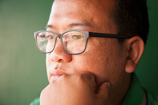 Indian man from Nagaland, India at home.  He wears a green collared shirt and glasses and a serious expression.  He stands near a green wall.  Portrait-style image of this man who is in his late 20s.  
