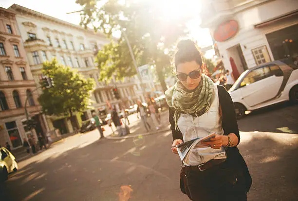Female tourist walking on the streets of Berlin's Kreuzberg district, looking at the map as she crosses the street in the sunset hours. She is wearing sunglasses and a scarf.