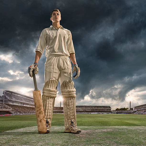 Cricket Batsman Hero A low angle portrait image of a professional cricket batsman dressed a full cricket whites, pads, gloves and leaning on cricket bat looking off camera into the distance. The player is standing on a cricket pitch in a generic floodlit outdoor cricket ground full of spectators under a dark stormy evening sky.  batsman photos stock pictures, royalty-free photos & images