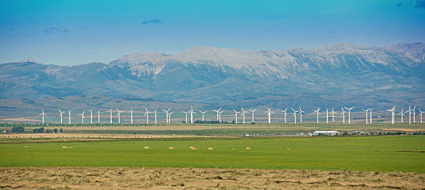 On The Southern Alberta, Canada,  prairie,  electricity producing wind turbines along a vast expanse of land with much of it in crops. The Rocky Mountains are in the background.