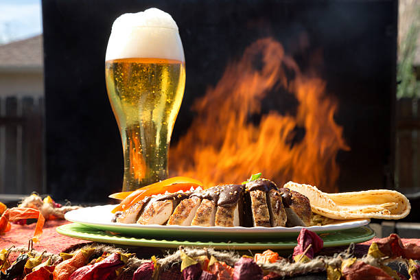 Rich mole sauce poured over sliced grilled chicken breast tacos with corn tortillas and a flaming barbecue grill in the background in a front or back yard with Mexican decorations in the foreground and a frosty glass of foamy beer to drink.