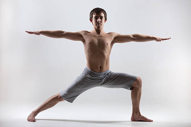 Sporty muscular young man working out, yoga, pilates, fitness training, doing lunge exercise, Warrior II posture, Virabhadrasana 2, gray background, low key shot
