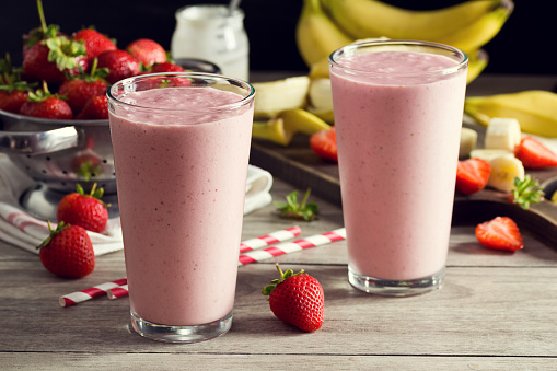 Two cold strawberry banana yogurt smoothies in pint glasses with striped straws. The berries and bananas are in the background in a colander and on a cutting board. A jar of yogurt is also in the background. Everything is on a faded wood picnic table.
