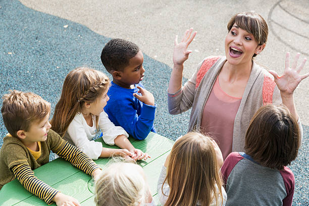 Teacher with preschool children having fun High angle view of a preschool teacher with a multi-ethnic group of children, 4 to 5 years old, sitting at a green table outdoors.  The preschoolers are looking at their teacher, listening to her tell a story.  She is animated, smiling with her mouth wide open and her hands raised. teacher classroom child education stock pictures, royalty-free photos & images