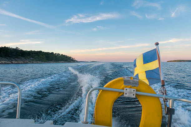 By boat in sunset, Stockholm Archipelago, Sweden. Wake of the boat on a wide expanse of sea with sunset in background. The silhuettes of island visible in the background, in the foreground yellow lifebuoy and Swedish flag. Summer, Clear sky.