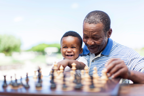 African grandfather sitting down with his grandson on his lap, teaching him how to play chess in Langebaan, Western Cape, South Africa