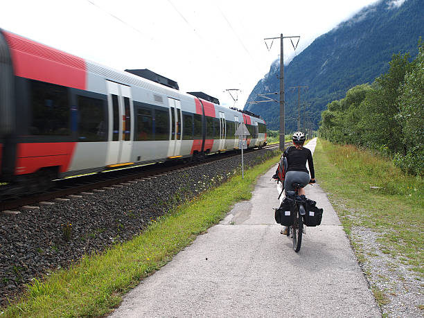 Back view of Biker riding on Drau cycle path near ruins of Aguntum in Tirol, Austria whiles the train in passing by. It is located in Valley of the Drava River near border of north Italy and Austria. One of nicest world Drau bike cycle paths ( Drauradweg R1) goes from Toblach trough Innichen in Italy, Weitlanbrunn and Villach in Austria and to Maribor in Slovenia. It is grey cloudy and rainy day...