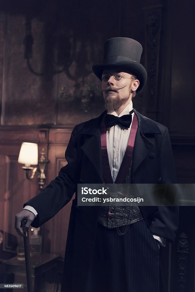 Vintage 1900 fashion man with beard and black hat. Vintage 1900 fashion man with beard and glasses. Standing in old wooden room.  Wearing a black dickens hat. Holding walking stick. Men Stock Photo