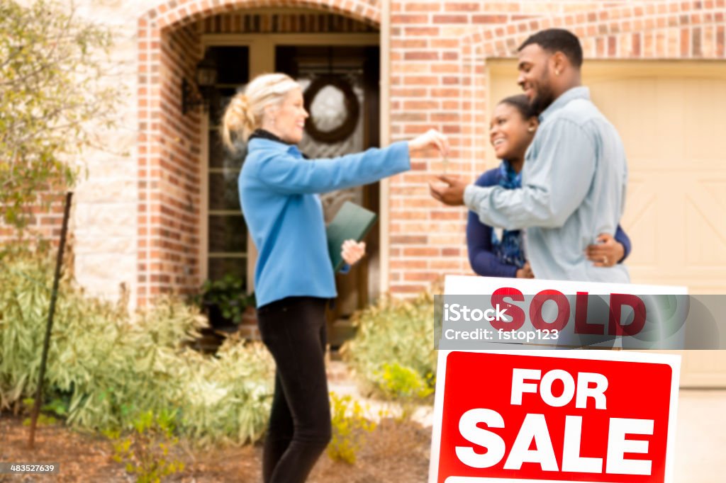 Real Estate: African descent couple buys home. Real Estate Agent gives key. African descent couple receives key from Real Estate Agent after purchasing a new home. 'Sold',  'For Sale' sign in foreground. Front yard view of this beautiful brick and stone home.  Selling Stock Photo