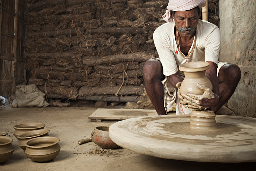 Day time image of traditional Indian potter busy in making clay pots with his skilled shaping hands on manual pottery wheel in his workshop. In the background are stacks of dried cow-dung used in baking the pottery. Horizontal composition with copy space and selective focus.