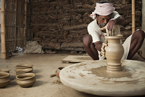 Day time image of traditional Indian potter busy in making clay pots with his skilled shaping hands on manual pottery wheel in his workshop. In the background are stacks of dried cow-dung used in baking the pottery. Horizontal composition with copy space and selective focus.