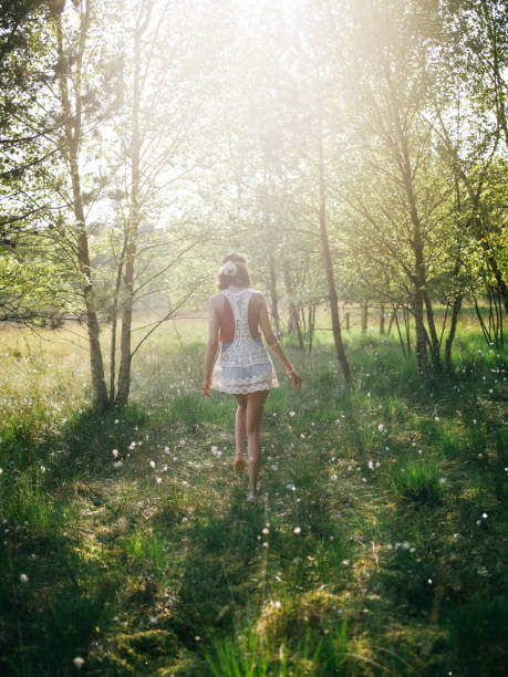 Full length rearview shot of a hippie girl in a vintage white lace top walking through lush green grass and trees in a summer park