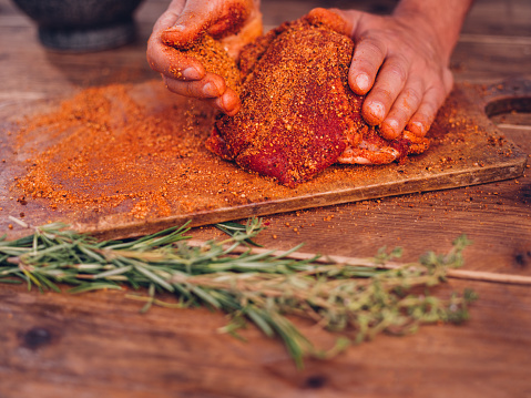 Low angle shot of some spicy seasoning being rubbed into a piece of raw pork on a wooden surface with herbs in the foreground