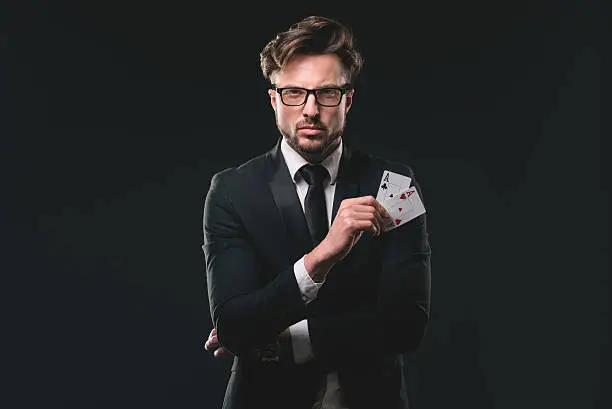 Studio portrait of young man with glasses wearing suite and necktie and holding aces of clubs and hearts. Black background.