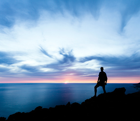 Man hiking silhouette in mountains, sunset and ocean. Male hiker with backpack on top of mountain looking at beautiful night landscape.