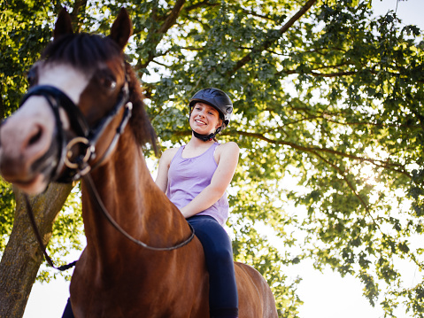 Teen girl horserider looking lovingly at her beautiful horse while riding it outdoors under lush green leafy trees