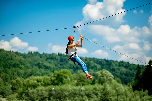 Young woman in casual wearing with red helmet riding on a zip line in the mountains. Active kind of recreation