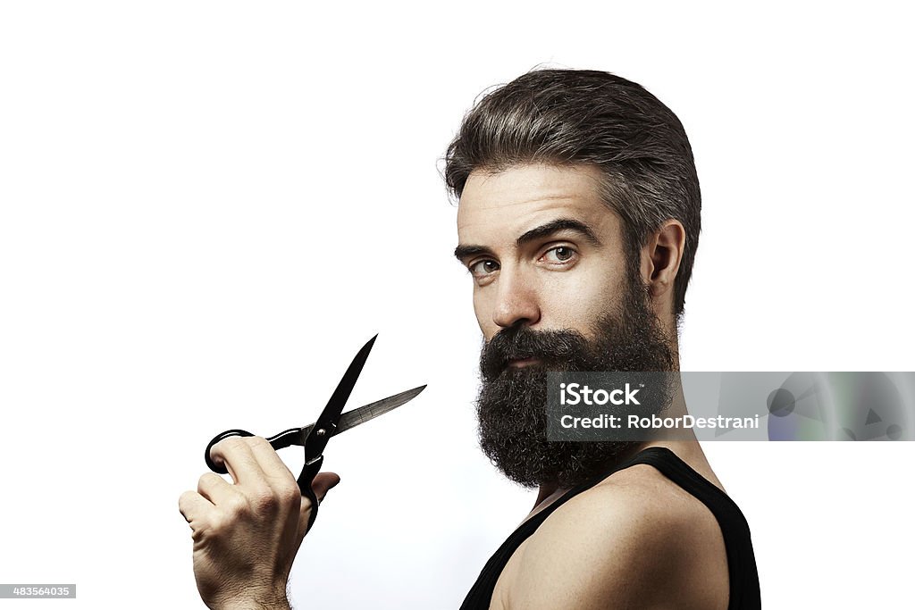 Bearded man holding scissors and wearing undershirt on white background Young man wearing undershirt and holding scissors photographed against a white background and looking at camera Men Stock Photo