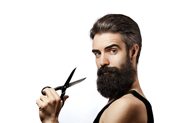 Bearded man holding scissors and wearing undershirt on white background Young man wearing undershirt and holding scissors photographed against a white background and looking at camera bushy stock pictures, royalty-free photos & images