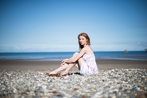 Portrait of a young redhead woman sitting on a pebble beach in the British Isles. She is wearing a summer dress with the horizon of the sea behind her.