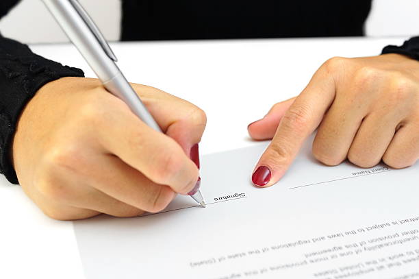 Woman's hands signing contract document A pair of woman's hands are over a white piece of paper on a white table. Her right hand is holding a shiny gray pen with its point touching the page above a line marked "Signature." Her fingernails are red. The index finger on her left hand is points to the signature line. The paper is upside down to the viewer. The woman is wearing a long-sleeved black shirt. A section of her torso is visible in the background past the edge of the table. The rest of the woman is not visible. witness signature stock pictures, royalty-free photos & images
