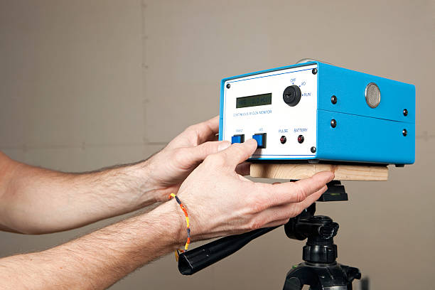 Hands programming a continuous radon monitor mounted to a tripod in an unfinished house basement. A house is commonly tested for radon with a single use test kit. If initial readings are high or remediation work has begun a system like this isz used to determine a more accurate measurement over time. An added benefit of this unit is real time measurement. The display screen reads Rate 2.3 pCi/l  (picocuries per liter), the curie is a unit of radioactivity. The background is unfinished drywall with seams and screws.