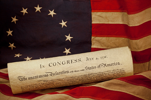 United States of America Declaration of Independence dated July 4, 1776 lying on an American flag. The flag, popularly attributed to Betsy Ross, was designed during the American Revolutionary War and features 13 stars to represent the original 13 colonies.