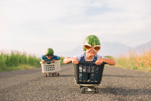 A young boy races his brother in a makeshift go-cart while wearing watermelon helmets and goggles. He is excited as he is winning the race.