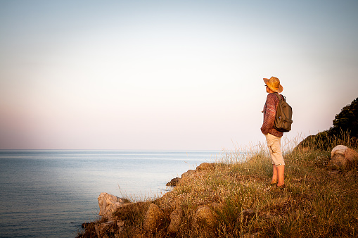 Woman with backpack standing on the top of the mountain in the sunset and enjoying the sea view