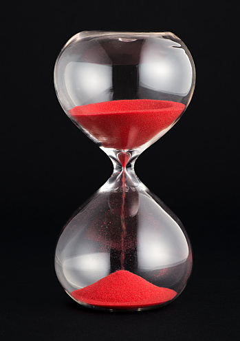 Hourglass with red sand running through the glass bulbs counting down the remaining time as it measures the passing minutes or hours, on a black background