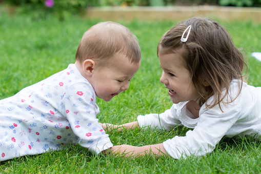 A baby and a toddler girl lying on the grass facing each other