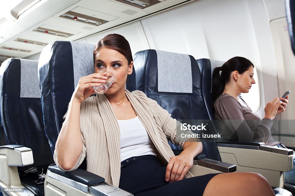 On the airplane Young woman sitting on the airplane and drinking water, while woman in the background using a smart phone. Airplane Stock Photo