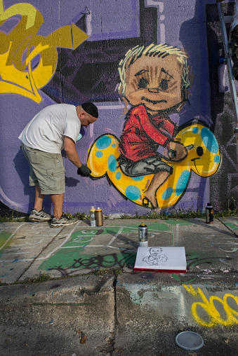 Miami, United States - December 5, 2012: During Art Basel week a graffiti artist paints a mural on a wall in Wynwood known as the Miami Wall of Fame. His sketch book is open on the sidewalk with an illustrated rough draft of the character he is painting.