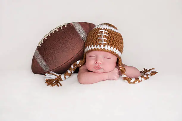 Portrait of a two week old, sleeping newborn baby boy. He is posed with an American football wearing a crocheted football hat.