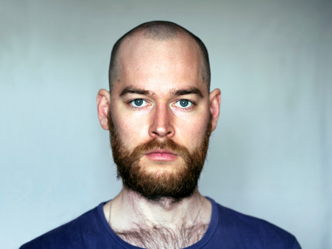 A man with a shaven head and big ginger beard looking at camera. Grey background.
