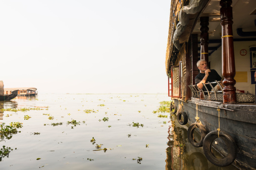 A senior man on house boat eating ice cream and  sails down the river in backwaters  In Alappey, Kerala, India.  Reflection in water. Kerala state, with a large network of inland canals earning it the sobriquet 