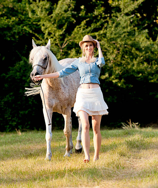 smiling young woman with straw hat walking with horse on meadow looking at the camera