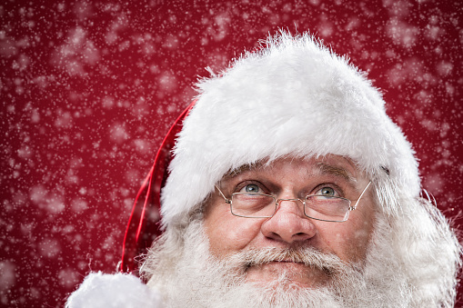 Closeup of the face of authentic Santa Claus with a real beard, looking up with a thoughtful little smile. Shot on a red background.