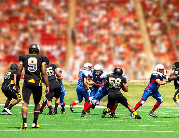Football team's running back carries ball. Defenders. Stadium fans. Field. Semi-professional football team's running back carries the football to make a play. Defenders try to tackle him. Football field with a stadium full of unrecognizable fans in background. defending sport photos stock pictures, royalty-free photos & images