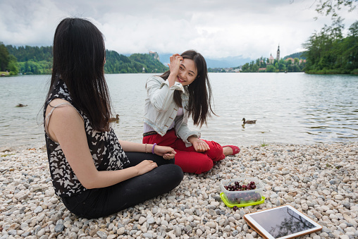 Two young Chinese women with dark long hair sitting on the pebbles on the Bled lake shore and eating cherries. One in red trousers is laughing. The other in black is turned towards the lake(rear view). Digital tablet and the cherry box on the floor. The castle and the island with a church in the background. Cloudy summer day, Bled, Slovenia, Europe. Nikon D800, full frame, XXXL.