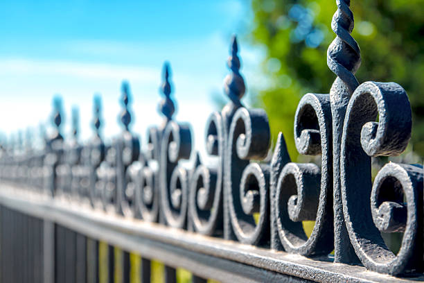 Horizontal composition photography of close-up in selective focus of iron fence, barrier, ornate with volute and curve. The black railing is taken in dimishing perspective on blurred blue sky and green tree background in sunny day of summer season.