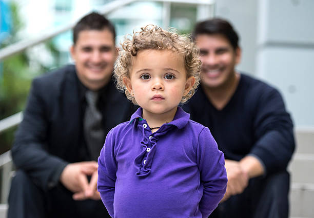 Beloved Child Two years old little girl posing with two smiling adult men in the background surrogacy stock pictures, royalty-free photos & images