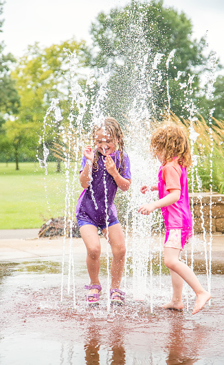 Two young girls (sisters) playing in a fountain at the splash park on a summer day. The older sister is in purple and is standing in the middle of the fountain as the water gets higher. She is about to jump out of the fountain as her younger sister, in pink, is just stepping into it. Taken on an overcast summer day.