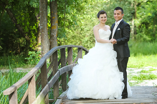 The bride and groom posing on a wooden bridge in nature, holding hands and looking at camera.