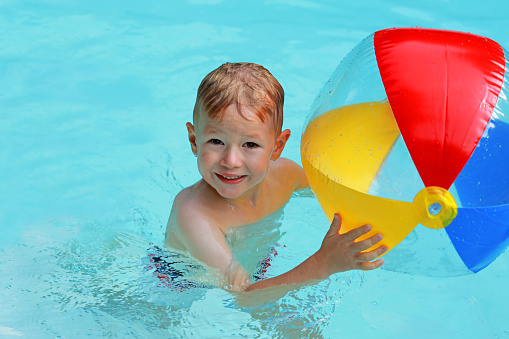 Little boy is smiling as he is holding a big colorful beach ball in an outdoor swimming pool in summer. This is a close-up picture of the swimming child and the ball in bright yellow, red, and blue colors.