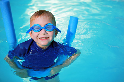 Little boy wearing a blue uv swimsuit and blue swimming goggles is smiling as he is learning to swim holding a noodle float in an outdoor swimming pool in summer. This is a close-up picture of the swimming child only.